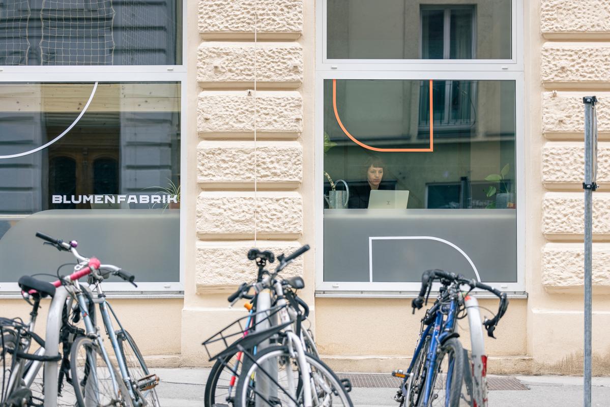 Hausfasade von außen, mit Rädern im Vordergrund. Im großen Fenster sitzt eine Frau und arbeitet an ihrem Laptop.