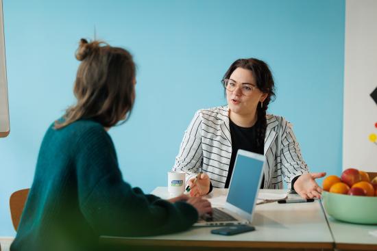 Zwei Frauen sitzen an einem Tisch uns sprechen miteinander. Vor ihnen befindet sich ein Obstkorb und ein Laptop.