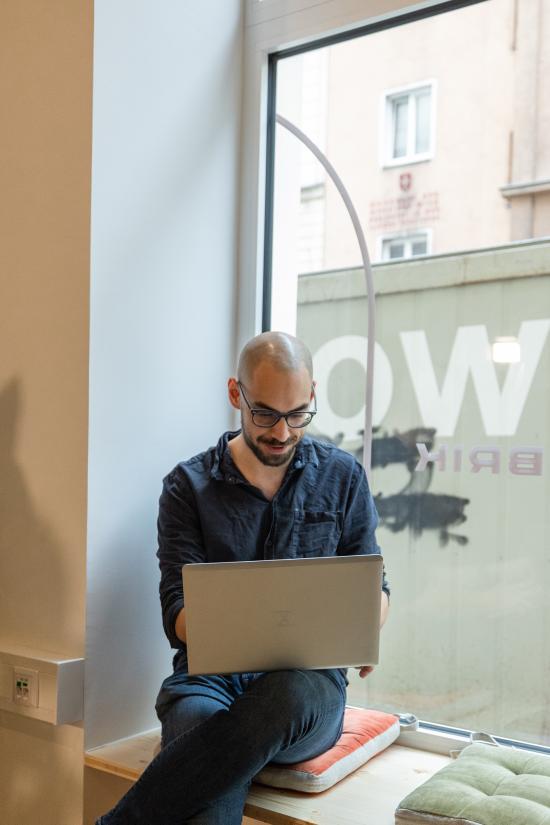 ein Mann mit Glatze und Brille sitzt an einer Fensterbank und hat einen Laptop auf seinem Schoß.