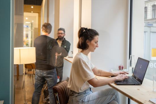 Frau sitzt an einer Fensterbank und arbeitet an einem Laptop. Im Hintergrund sieht man zwei Männer im Gespräch.