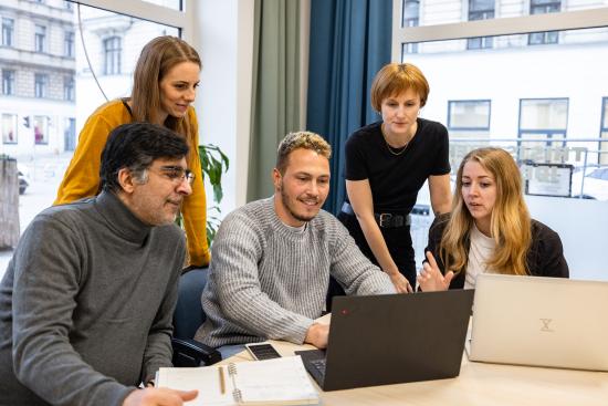 zwei Männer und eine Frau sitzen an einem Meeting Tisch, sie schauen auf einen Laptop-Bildschrim, dahinter stehen zwei weitere Frauen, die ebenfalls interessiert darauf schauen. 