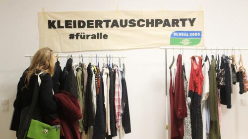 Frau begutachtet bei einer Kleiderstange verschiedene Kleidungsstücke. Über der Kleiderstange befindet sich ein Bunner worauf steht "Kleidertauschparty #füralle"